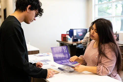 A student consults with a staff member at a front desk, reviewing brochures and information materials in a bright office setting.